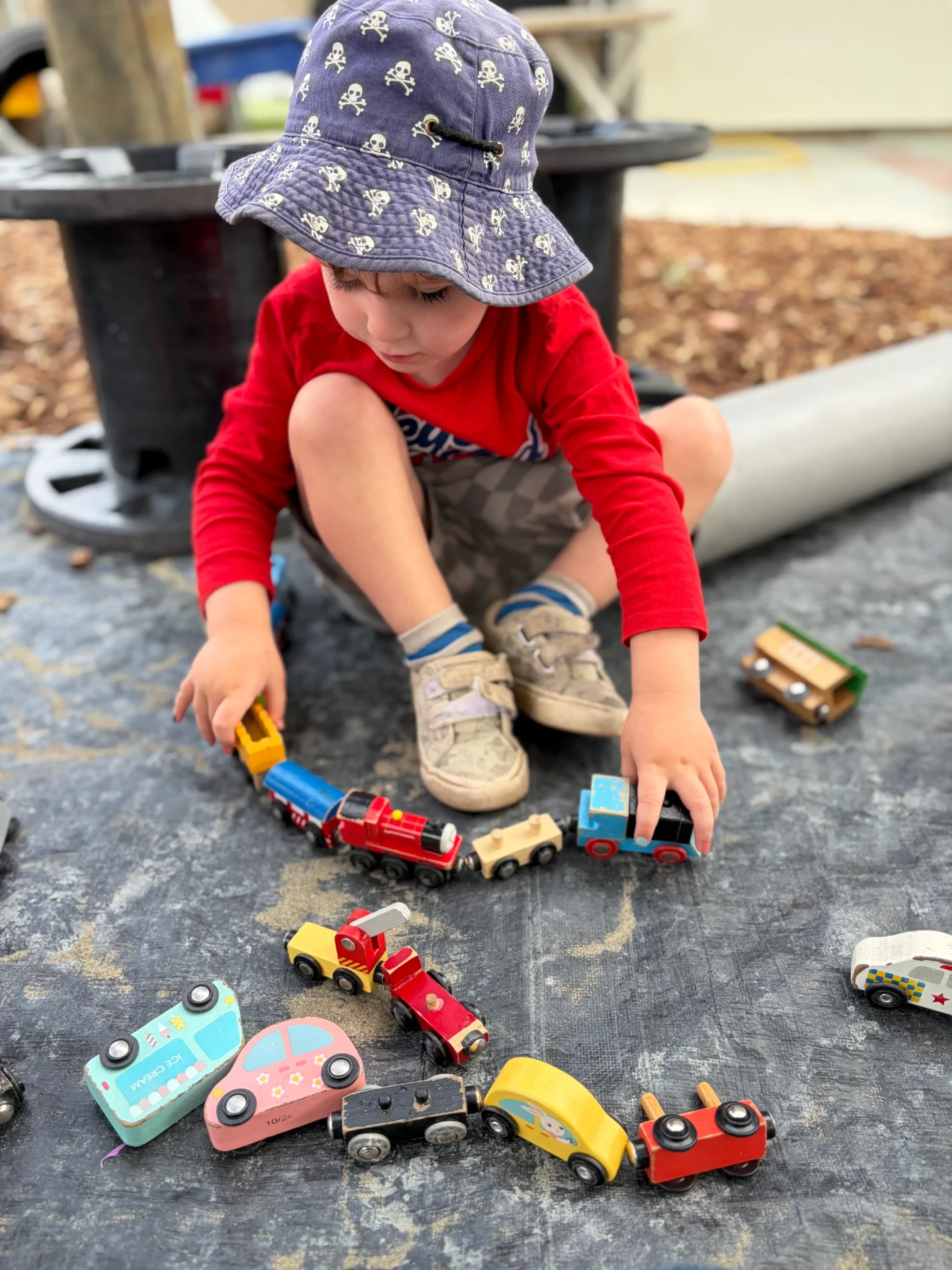 A toddler in a sun hat lining up wooden trains on the mat