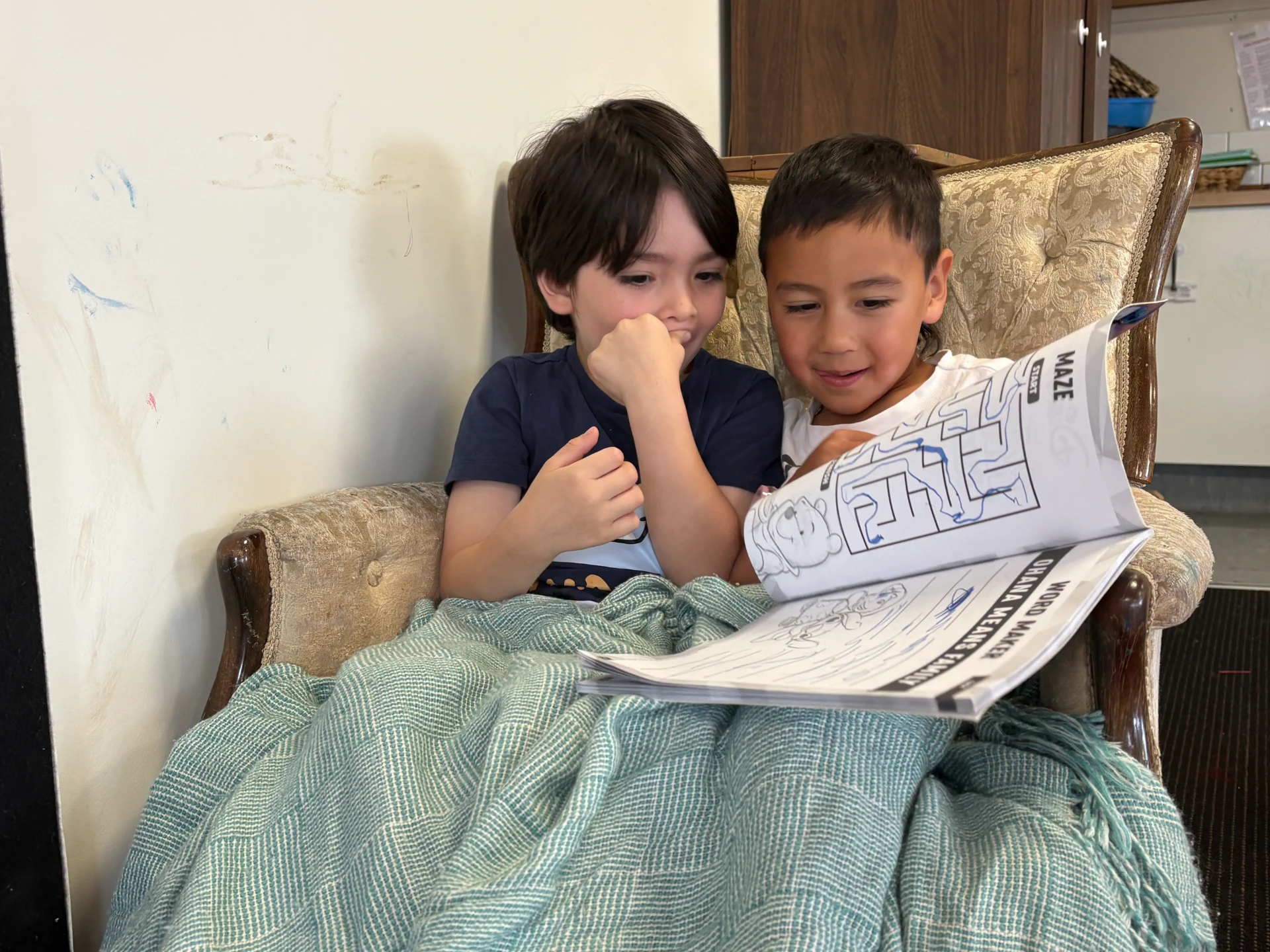Two preschoolers working through a maze book together under a blanket in the armchair