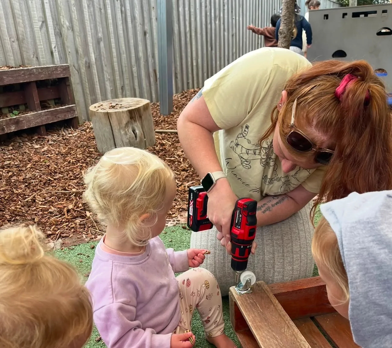 A kaiako using a cordless drill to fix a wooden trolley while tamariki watch closely