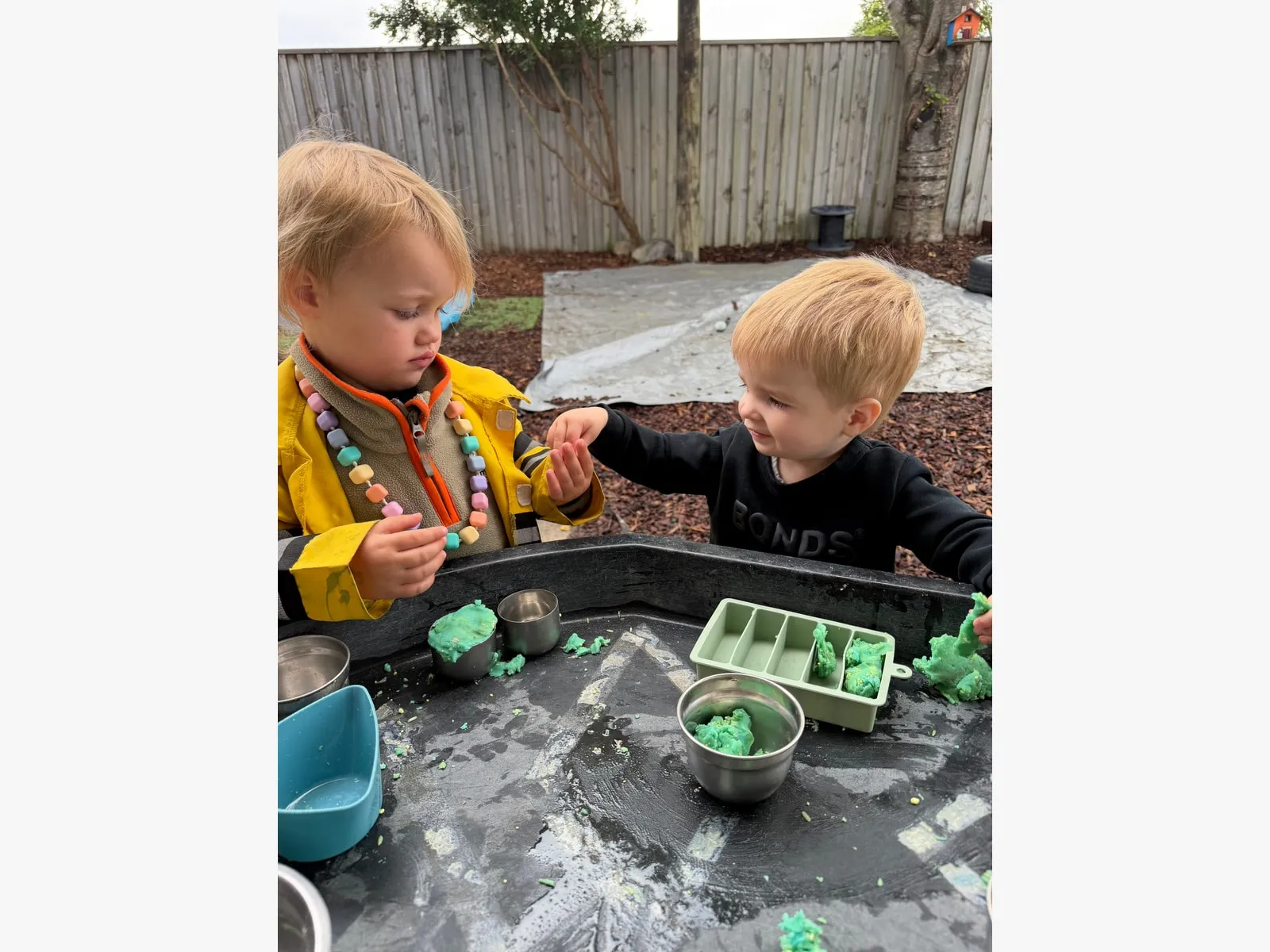 Two toddlers sharing scoops and moulds at the outdoor sensory table