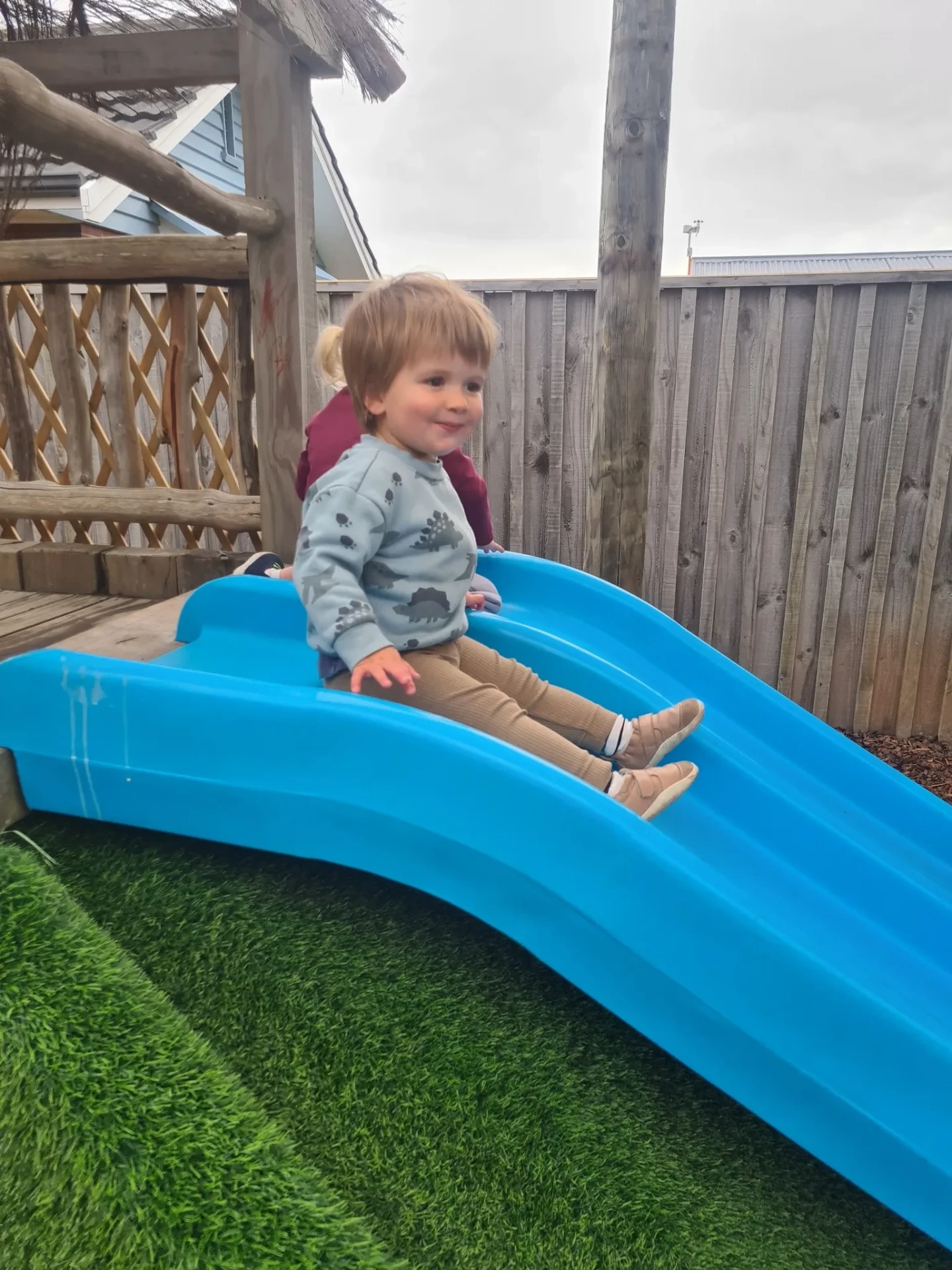 A smiling toddler sitting at the top of the blue slide outside