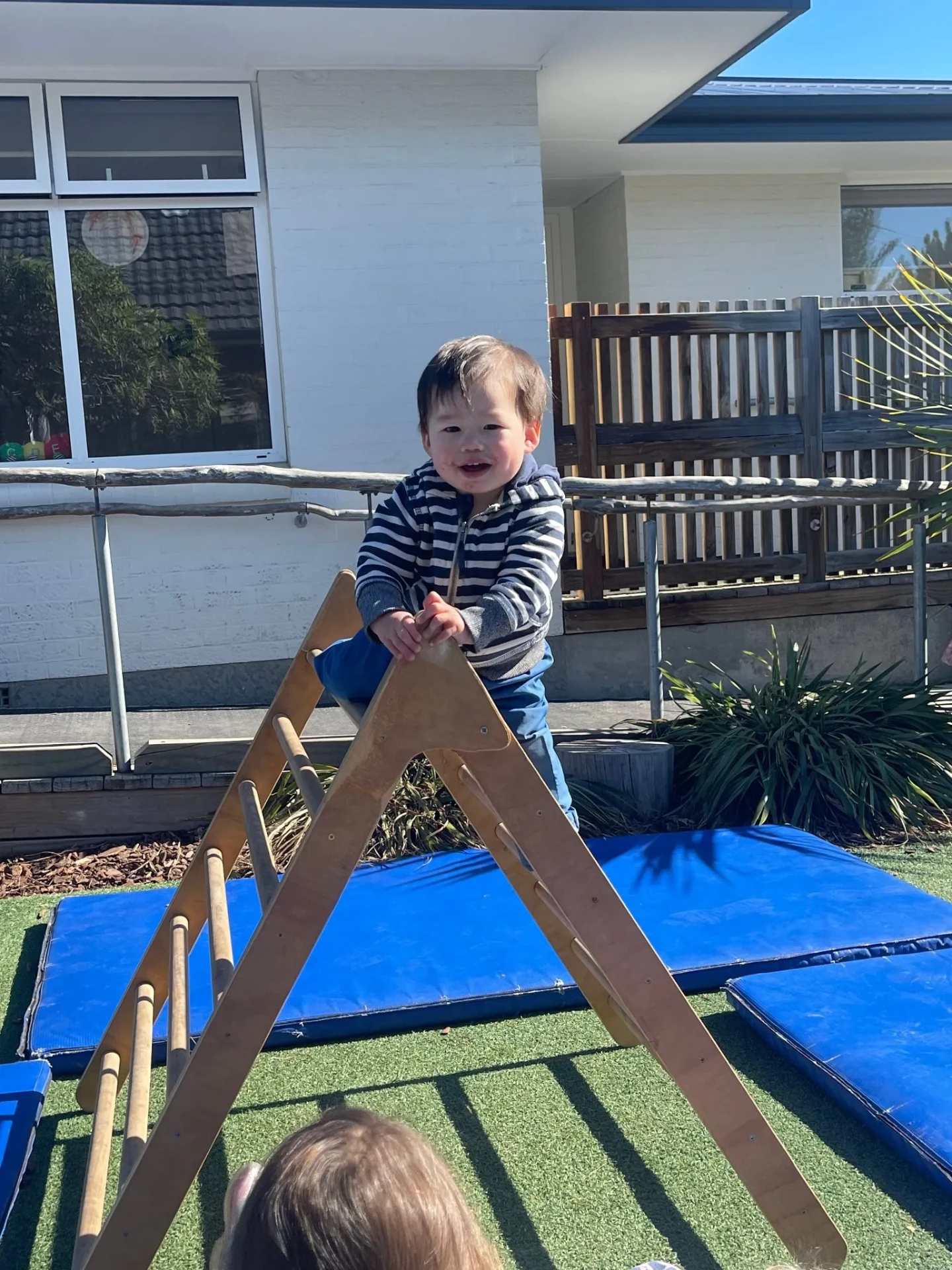 A toddler smiling at the top of a wooden climbing frame above soft mats