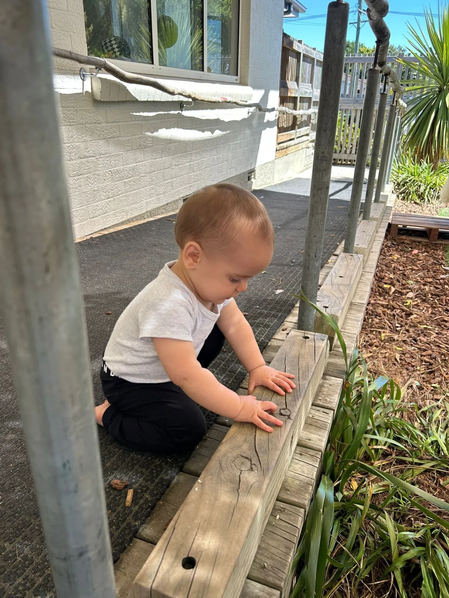 A baby sitting outside exploring a wooden garden edge in the sunshine