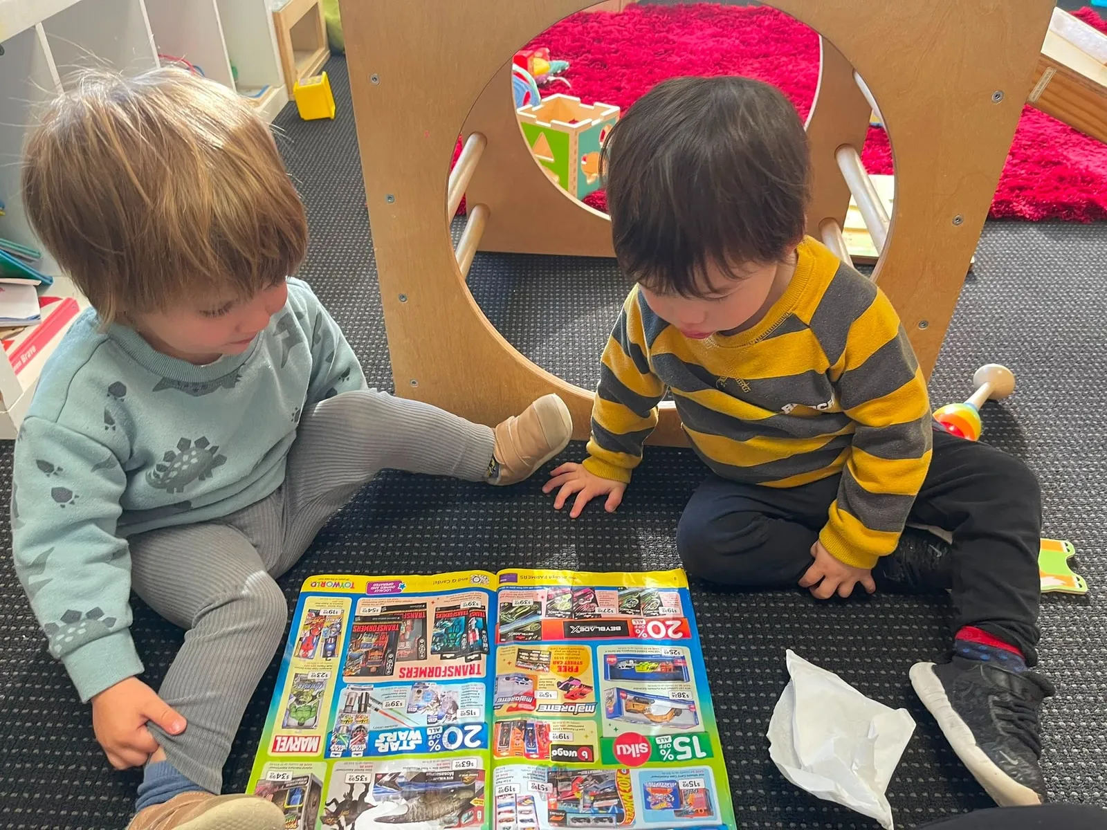 Two toddlers sitting on the floor turning the pages of a picture catalogue