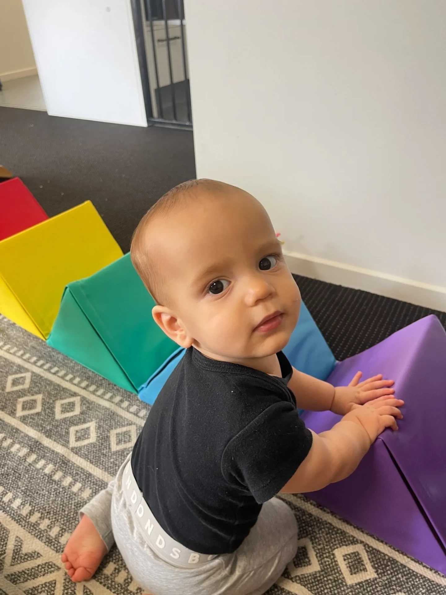 A baby pulling up on a soft play wedge and looking over their shoulder