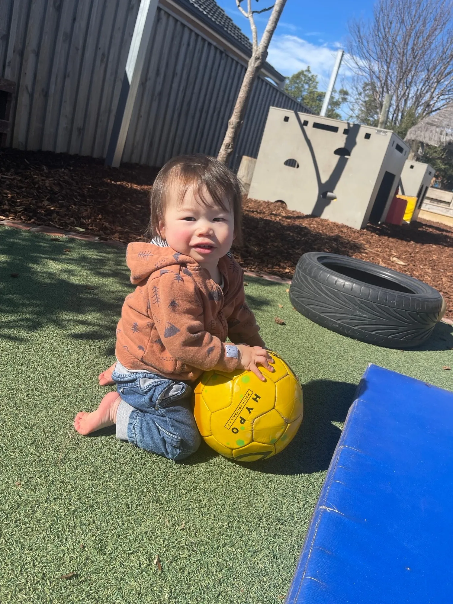 A toddler kneeling on the grass with a yellow ball outside